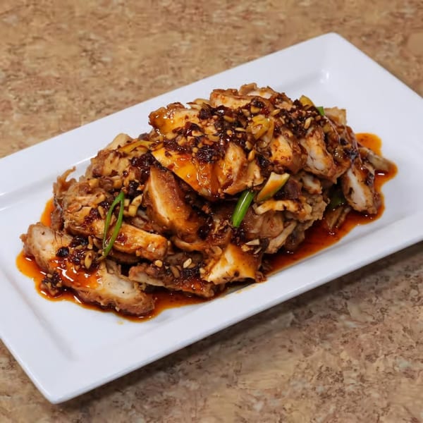 Sliced chicken with a dark, spicy sauce, peanuts, and green onions on a white rectangular plate.