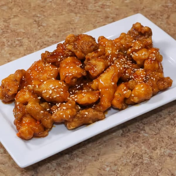 A white rectangular plate filled with glistening orange chicken pieces, sprinkled with white sesame seeds.